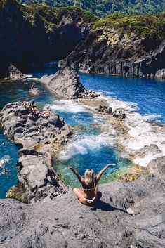 a woman sitting on top of a rocky cliff next to the ocean with her arms in the air