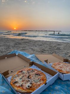 two pizzas sitting on top of a blue blanket next to the ocean at sunset