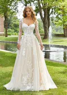 a woman in a wedding dress standing on the grass with trees and water behind her