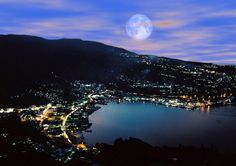 the city lights are lit up at night on the water's edge with mountains in the background