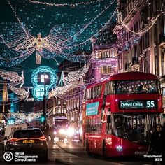 a red double decker bus driving down a street next to tall buildings covered in christmas lights