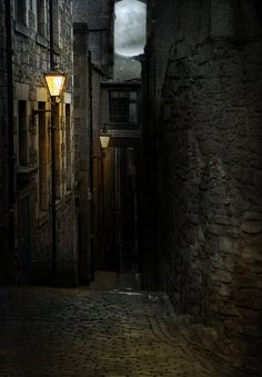 an alley way with stone walls and cobblestone flooring at night, lit by street lamps