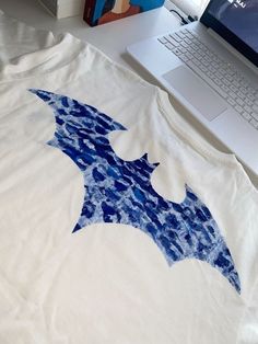 a white t - shirt with a blue bat on it sitting next to a laptop