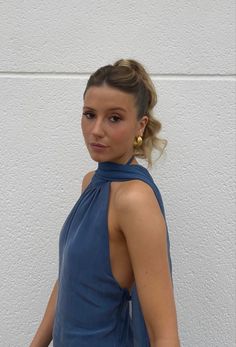 a woman standing in front of a white wall wearing a blue top and gold earrings