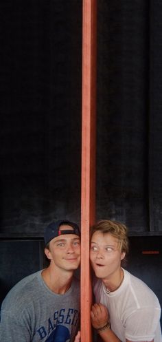 two men standing next to each other in front of a tall wooden pole with a clock on it's side