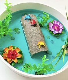 a bowl filled with water and plants on top of a blue surface, next to flowers