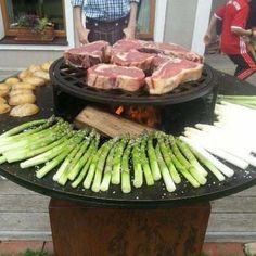 an outdoor grill with asparagus and steaks on it, in front of people