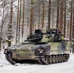 an army tank driving through the snow in front of some trees and pine trees with no leaves on it