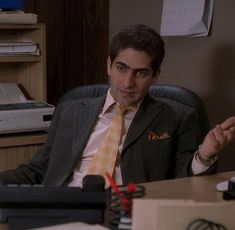 a man in a suit and tie sitting at a desk with his hand out to the side