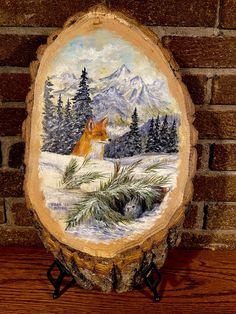 a painting of a fox in the snow on a wood slice with trees and mountains behind it