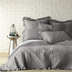 a bed with grey linens and pillows on it in front of a white brick wall