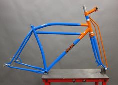 an orange and blue bike frame sitting on top of a red table next to a gray wall