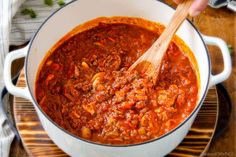 a wooden spoon in a white pot filled with chili and meat sauce on top of a cutting board