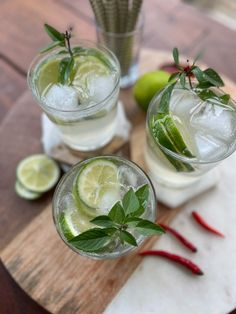 two glasses filled with ice and limes on top of a cutting board