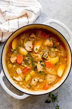 chicken and vegetable soup in a white pot on a gray surface with a towel next to it