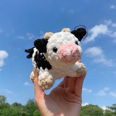 a hand holding a small stuffed cow in front of a blue sky with clouds and trees