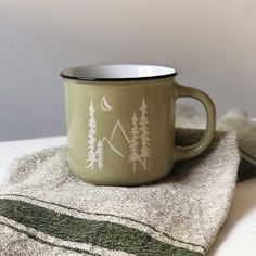 a green coffee mug sitting on top of a towel next to a cup with the moon and trees painted on it