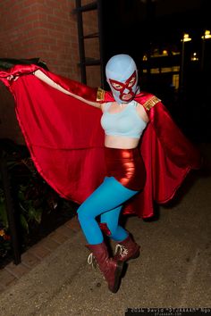 a woman dressed in costume poses with a red cape over her shoulders and blue tights