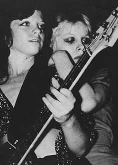 black and white photograph of two women playing bass guitars in front of a man with his arm around the woman's neck