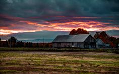 an old barn sits in the middle of a field as the sun sets behind it