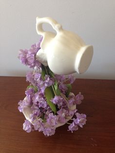 a white vase with purple flowers in it on a wooden table next to a wall