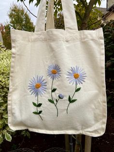 a white tote bag with blue flowers painted on the front and side, hanging from a tree