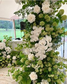 white flowers and greenery are arranged in a vase on a wooden table under a tent