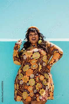 a woman standing in front of a blue wall wearing a brown and yellow dress with flowers on it