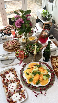 a table full of food and drinks with flowers on the table top, in front of a large window