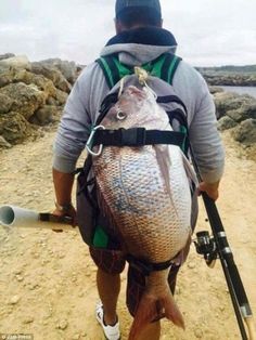 a man walking down a dirt road while holding a fish in his back pack and fishing pole