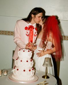 a man and woman kissing in front of a white cake with red flowers on it