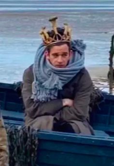 a man wearing a crown sitting in a blue boat on the beach next to the ocean