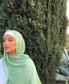 a woman wearing a green hijab standing in front of a tree and bushes