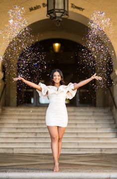 a woman in a white dress standing on steps with her arms outstretched and confetti falling from the sky