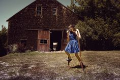 a woman standing in front of a barn with a backpack on her back and wearing a blue dress