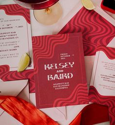 the wedding stationery is laid out on top of red and white paper, with a glass of wine next to it