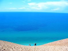 two people are walking on the beach near the ocean and sand dunes with blue water in the background