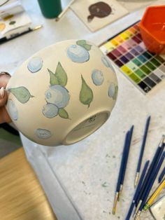 a person holding up a vase with blueberries on it and paintbrushes nearby