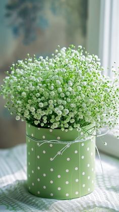 small white flowers in a green polka dot vase on a tablecloth with a window behind it