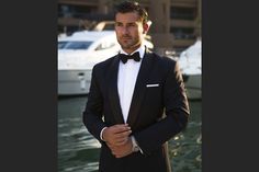 a man in a tuxedo standing next to some water with boats behind him