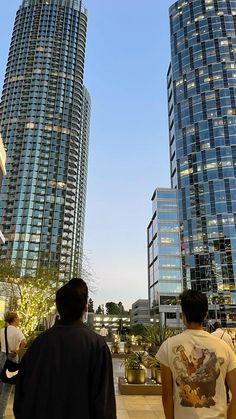 two men are standing in front of some very high rise buildings and looking up at the sky