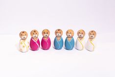 five small wooden dolls are lined up in a row on a white surface, one is wearing a blue dress and the other has a pink robe