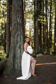 a pregnant woman in a white dress sitting on a bench next to a large tree