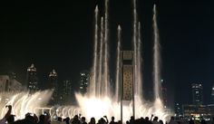 people are standing in front of a fountain at night with water shooting up from it