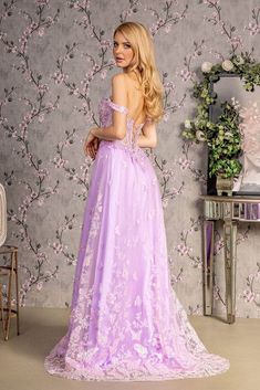 a woman standing in front of a floral wallpaper wearing a lavender colored gown with an open back