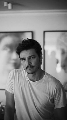 black and white photograph of man with mustache in front of two framed pictures on wall