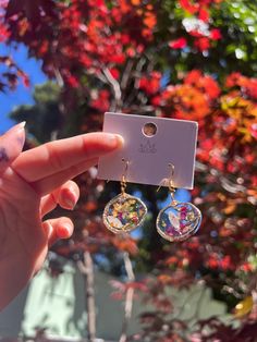 a person holding up a pair of earrings in front of a tree with red leaves