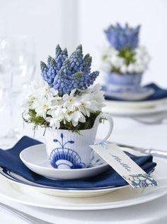 blue and white flowers are in a teacup on a table set for two with place settings