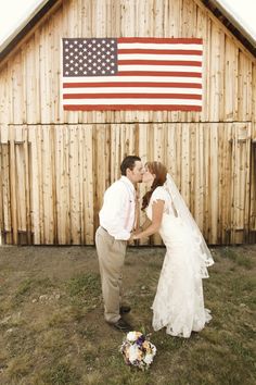 a bride and groom kissing in front of a barn with an american flag on it