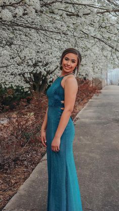 a woman in a long blue dress standing on a sidewalk next to some white flowers